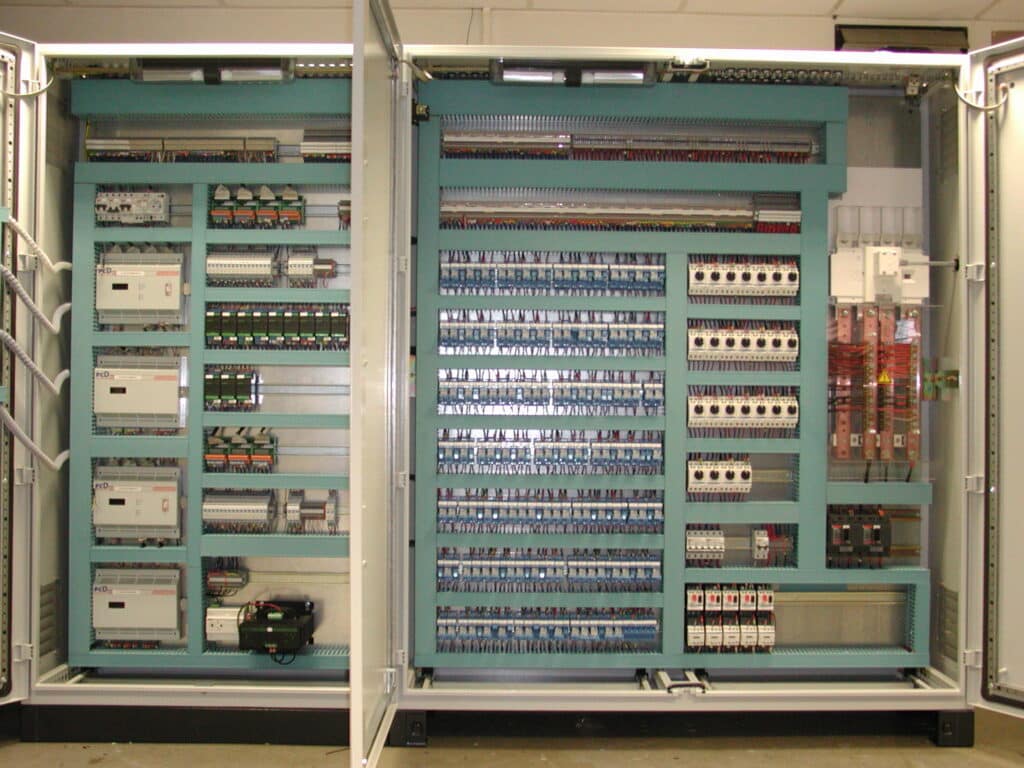 Intérieur d'une armoire électrique industrielle ouverte, avec PLCs 'pCD', câblage complexe, disjoncteurs et borniers.