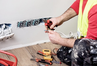 Électricien en gilet jaune installe des prises électriques murales. Il manipule des fils avec des pinces, des outils sont au sol.