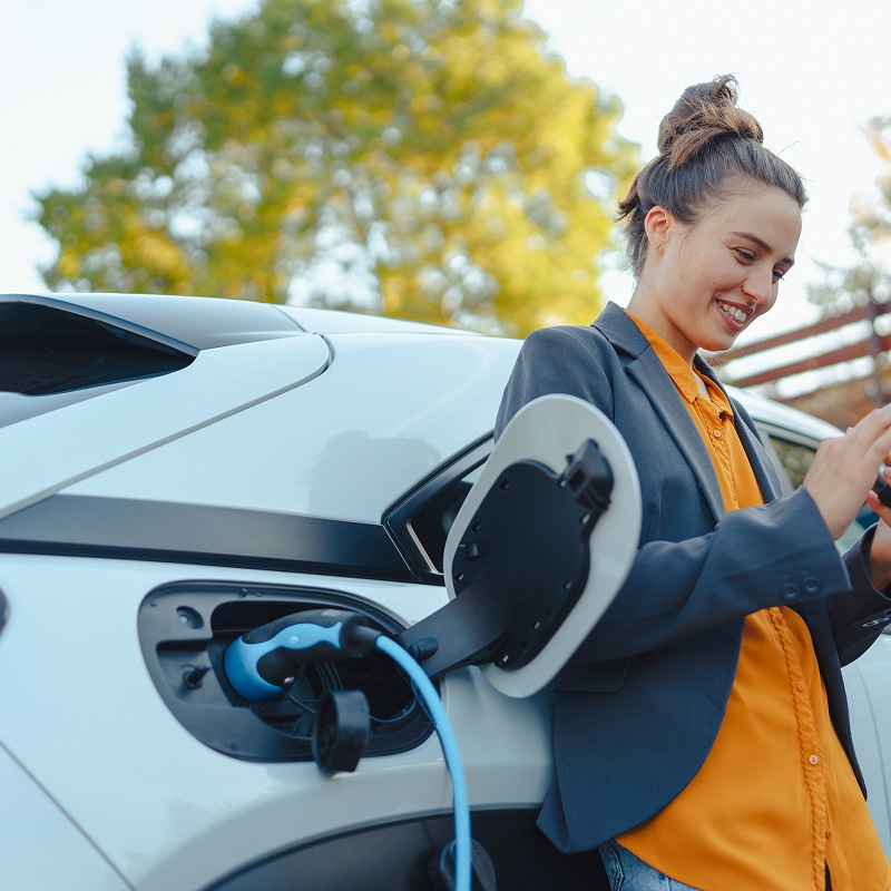 Femme souriante rechargeant sa voiture électrique blanche avec un câble bleu tout en regardant son téléphone. Arbres verts en arrière-plan.