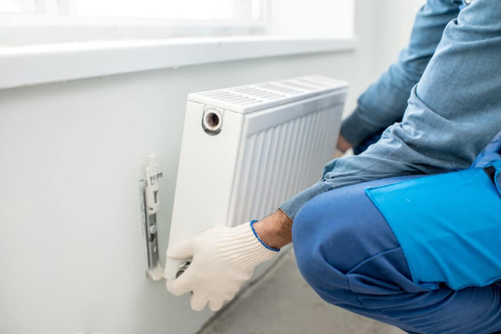 Un installateur en combinaison bleue et gants blancs fixe un radiateur de chauffage blanc au mur sous une fenêtre.