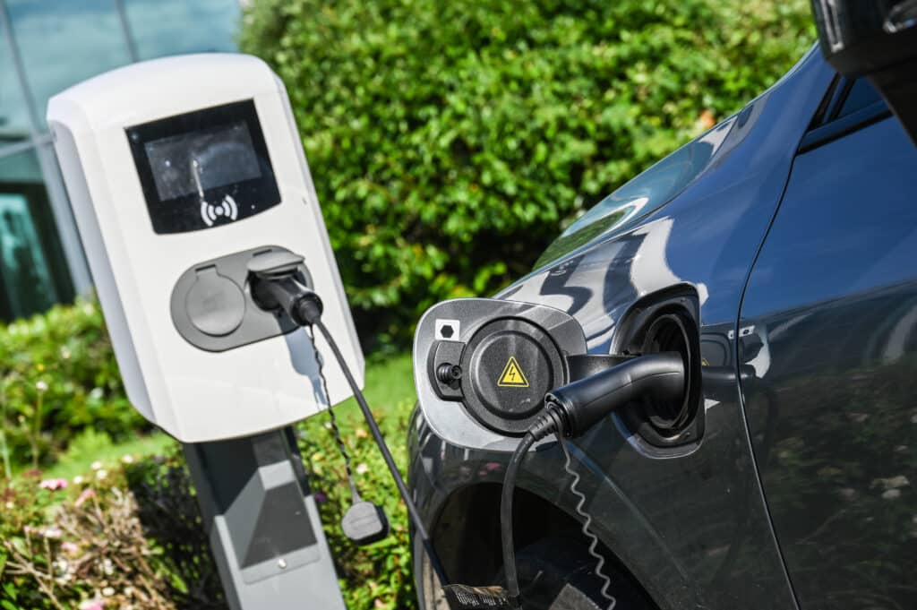 Voiture électrique grise en charge sur une borne blanche, câble branché. Symbole éclair sur le port du véhicule. Arrière-plan végétal flou.