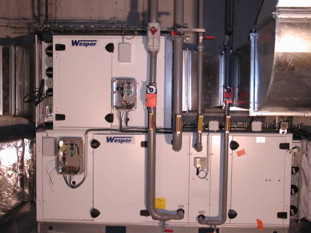 Unité de traitement d'air modulaire Wesper en salle mécanique. Panneaux blancs, tuyaux gris, vannes et conduits électriques. Connectée à des gaines métalliques.